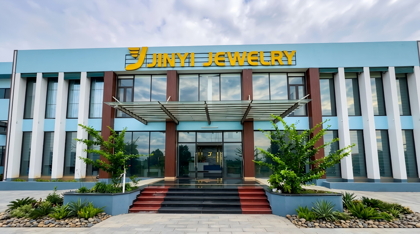 A centered, exterior photograph of the modern JINYI JEWELRY manufacturing facility, referenced in the file 'JIN YI.jpg'. The two-story building features a light blue facade with white columns and large floor-to-ceiling windows. The prominent, yellow three-dimensional logo and text "JINYI JEWELRY" are mounted above the glass entrance, which is covered by a glass canopy. The entrance is approached by a dark granite staircase with red-highlighted risers. The foreground is professionally landscaped with diverse green plants and river rocks, all under a partly cloudy sky.
